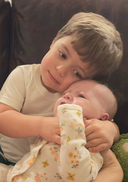 Baby Clara is meeting her older brother. He is holding her and hugging her on their couch at home.