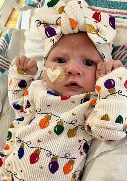 Baby Clara is in the NICU, dressed in her holiday lights onesie and matching headband bow