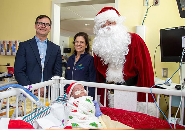 Baby Thomas is sleeping in his NICU bed with a Santa hat and blanket on. Behind him stands Santa Clause, and two from his care team.